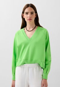 Tricou verde cu decolteu în V, mâneci lungi, man�șete cu nervuri și un tiv scurtat. Textură netedă, design casual, asociat cu pantaloni de culoare deschisă.