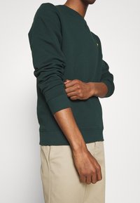 Sudadera verde bosque con cuello redondo, puños y dobladillo acanalados. Diseño simple con un pequeño logo en el pecho. Tela suave y texturizada.