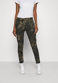 Pantalon à motif camouflage vert foncé avec des dessins de feuilles, doté de poches zippées et d'ornements texturés. Matière en mélange de coton.
