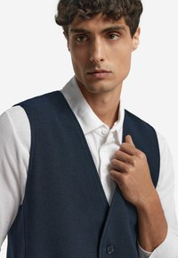 Gilet blu navy in tessuto tessuto, con design a v, chiusura con bottoni, indossato sopra una camicia bianca con colletto.