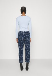 Personne avec des cheveux tressés portant un haut à manches longues bleu clair et un jean foncé à motifs avec des bottines noires, se tenant devant un fond blanc.