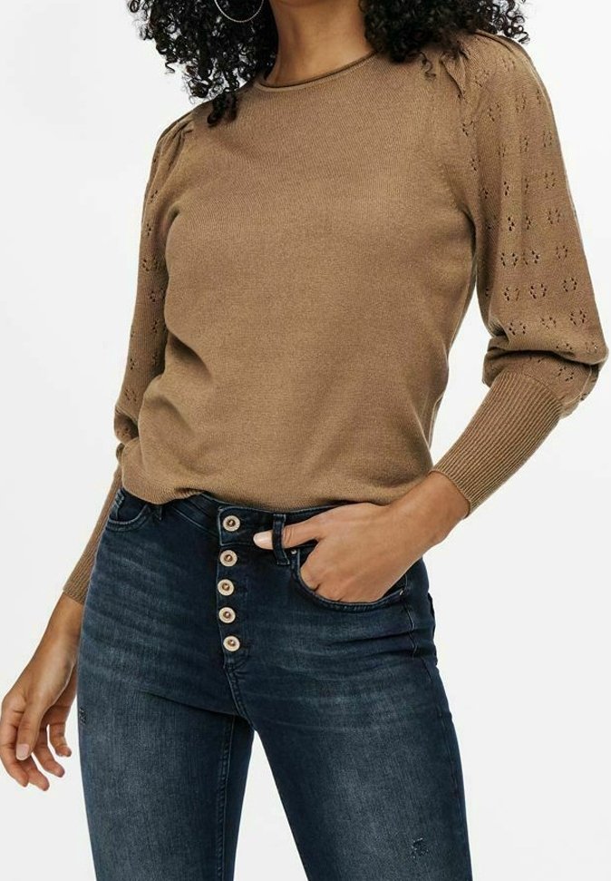 Femme portant un pull en tricot marron avec des manches bouffantes à motifs et un jean taille haute bleu foncé avec cinq boutons visibles.