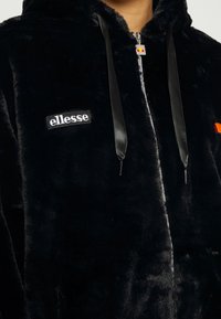 Sort fleece zip-hættejakke med en blank satin-snøre, der har et broderet logo-patch og et lille flerfarvet emblem på brystet.