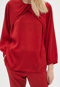 Czerwona bluzka z długim rękawem i skręconym detalem przy dekolcie, z elastycznymi mankietami, wykonana z gładkiego materiału. Połączona z pasującymi czerwonymi spodniami.