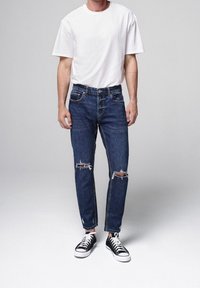 Man wearing a white t-shirt, ripped blue jeans, and black low-top sneakers standing against a plain gray background.