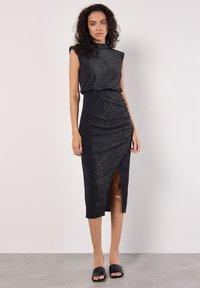 Vestido negro sin mangas con una tela texturizada y brillante. Presenta un escote alto, abertura lateral y detalles de fruncido en la cintura.