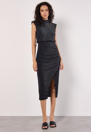 Vestido negro sin mangas con una tela texturizada y brillante. Presenta un escote alto, abertura lateral y detalles de fruncido en la cintura.