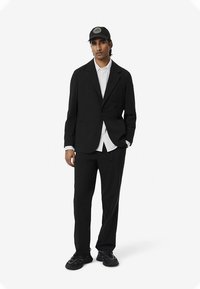 Férfi fekete öltöny szabott zakóval és nadrággal, fehér inggel rétegezve, fekete sapkával, amelyen logó található, és fekete sportcipővel párosítva.