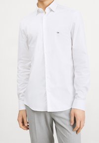 Man i en vit skjorta med knappar och "CK"-logotyp på bröstet, tillsammans med ljusgrå byxor, står med armarna avslappnat vid sidorna.