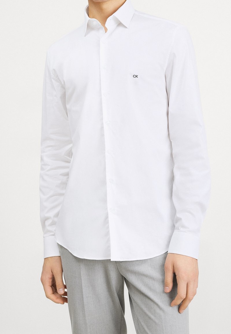 Man i en vit skjorta med knappar och "CK"-logotyp på bröstet, tillsammans med ljusgrå byxor, står med armarna avslappnat vid sidorna.