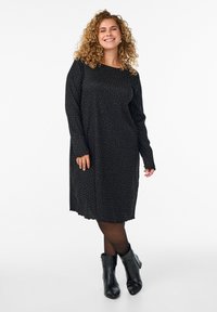 Robe noire à manches longues ornée de pois blancs, avec un col rond et un ourlet festonné, portée avec des bottines noires.