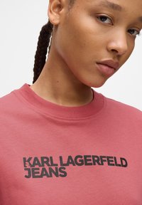 Raudonas medvilninis marškinėlių suapvalintu kaklu dizainas, kuriame yra juodas spaudinys "KARL LAGERFELD JEANS" per krūtinę; lygi tekstūra, laisvas siluetas.
