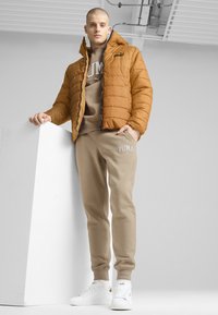 Giubbotto imbottito marrone con cappuccio, abbinato a pantaloni della tuta beige con logo e scarpe da ginnastica bianche. Texture lisce e design casual.