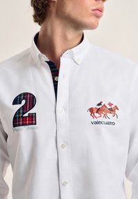 Camisa blanca de botones con un número "2" a cuadros y un diseño bordado de jinetes. Cuello forrado con tela a rayas.