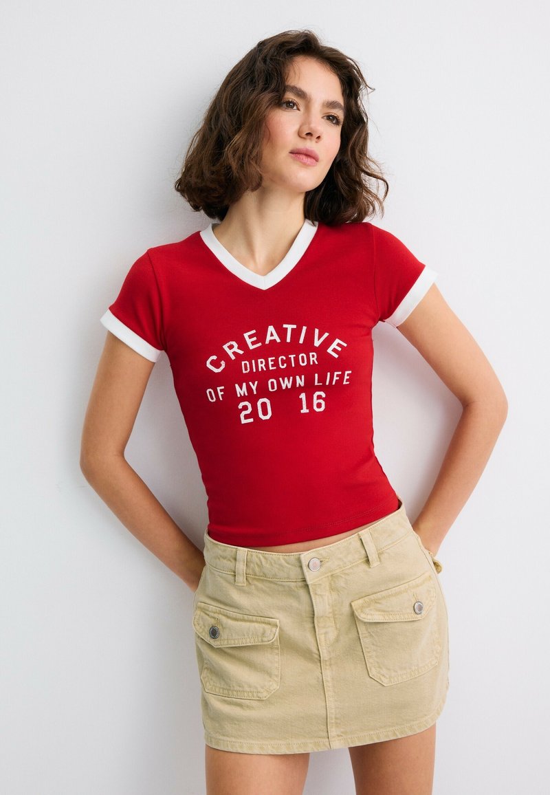 Jeune femme aux cheveux courts et bouclés, portant un t-shirt rouge avec un texte blanc et une jupe beige, debout sur un fond blanc.