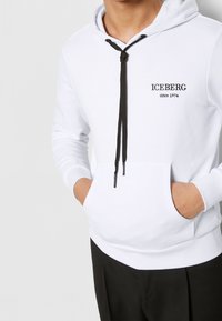 Felpa bianca con cappuccio, dotata di lacci neri e logo. Presenta una tasca frontale e polsini a coste. Realizzata in materiale morbido, adatta per un look casual.