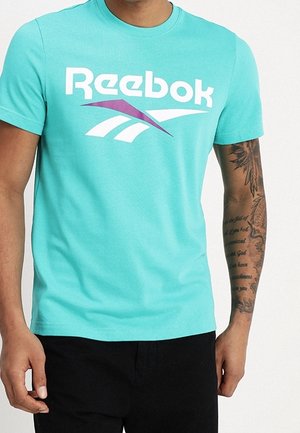 Mężczyzna w turkusowej koszulce Reebok i czarnych spodniach, prezentujący prawy, wytatuowany przedramię z tekstem i wzorem.