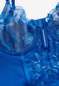 Mėlynas nėriniuotas liemenėlė su gėlėta siuvinėjimu, permatomu viršutiniu sluoksniu ir vientiso audinio pagrindu. Matomi reguliuojami dirželiai ir užsegimo kabliukas.