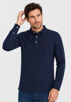 Hombre con cabello corto y rizado que lleva una camisa polo azul marino de manga larga y vaqueros, tocándose un lado de la cabeza y mirando hacia la derecha.
