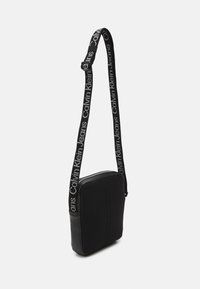 Borsa a tracolla nera con una parte anteriore in tessuto a rete strutturata, lati in pelle liscia e tracolla regolabile con il testo ripetuto "Calvin Klein Jeans".