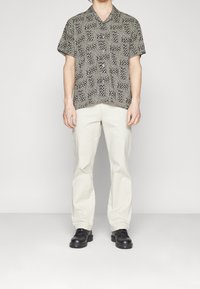 Košile s krátkým rukávem s černobílým vzorem, v kombinaci s světle béžovými rovnými kalhotami a černými botami, stojící na neutrálním pozadí.