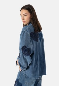 Denimjakke med mørkeblå paljett-hjertepatcher på skuldrene og ermene, med avslappet passform og frynsete kantdetaljer.