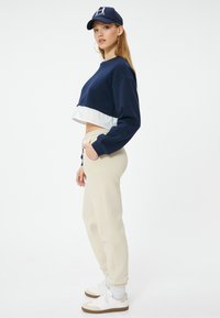 Marineblaues, verkürztes Sweatshirt mit weißen Akzenten, beigefarbene Jogginghosen und weiße Sneakers mit braunen Details. Accessoires sind eine marineblaue Mütze und Creolen.