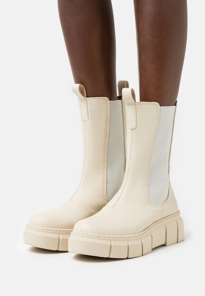 Bottes mi-mollet de couleur crème avec semelles épaisses portées sur des jambes à peau foncée, dotées de panneaux latéraux élastiques et de languettes d'extraction sur fond blanc.