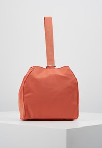 Kwadratowa torba z koralowego materiału z szerokim uchwytem przymocowanym u góry, wyeksponowana na białej powierzchni.