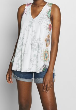 Nainen pukeutunut hihattomaan valkoiseen kukkakuvioiseen toppiin, jossa on värikkäitä pyöreitä kuvioita, ja sinisiin farkkushortseihin, joiden helmat on kääritty, seisoo yksinkertaisen taustan edessä.