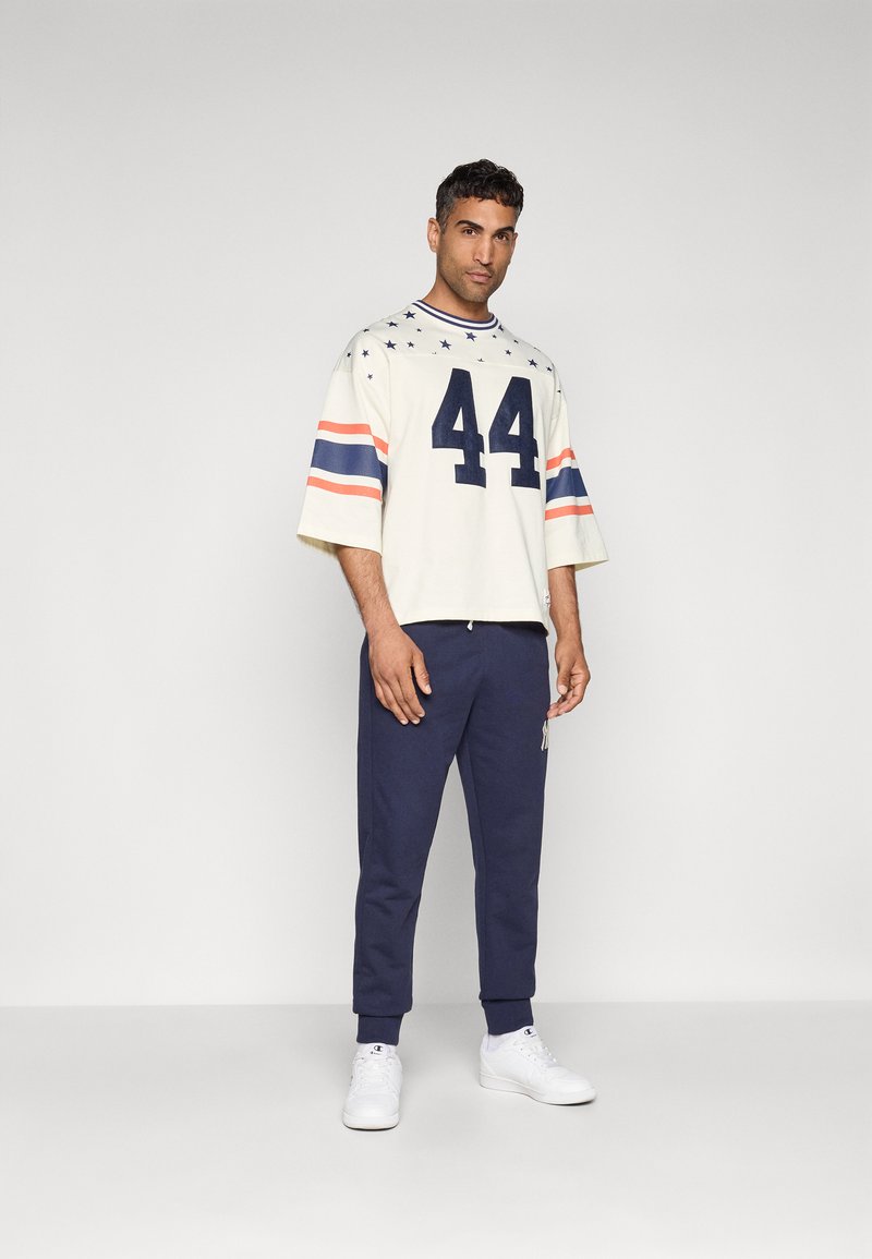 Crème oversized shirt met marineblauwe en oranje strepen, nummer 44, en sterrenpatroon; gecombineerd met marineblauwe sweatpants en witte sneakers.