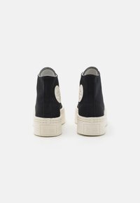 Black canvas ankle shoes with white rubber soles, featuring a round logo patch and stitched detailing along the edges.