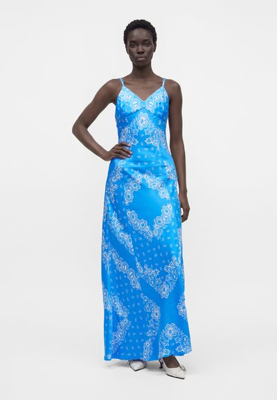 Femme grande aux cheveux courts portant une robe longue bleu sans manches avec des motifs floraux blancs et des talons blancs pointus, debout avec une main sur la hanche.
