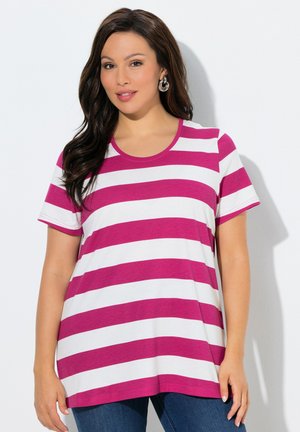 Frau mit langen dunklen Haaren, die silberne Creolen und ein kurzärmeliges T-Shirt mit pink-weißen horizontalen Streifen trägt, steht vor einer weißen Wand.