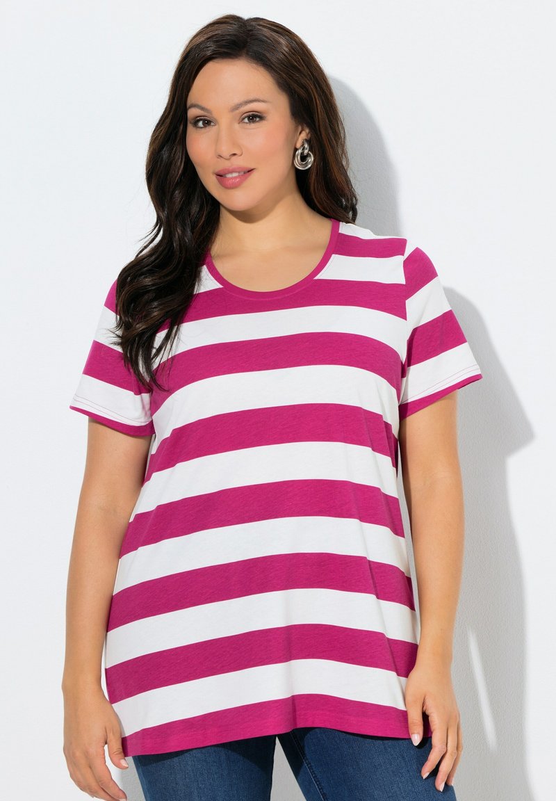 Donna con capelli lunghi e scuri che indossa orecchini a cerchio argentati e una T-shirt a maniche corte con righe orizzontali rosa e bianche, in piedi contro un muro bianco.