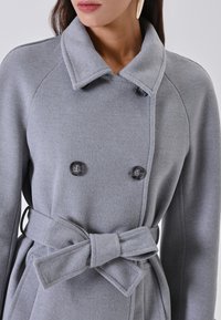 Cappotto grigio doppiopetto in tessuto testurizzato, con collo ampio e una cintura abbinata annodata a fiocco in vita. Bottoni neri come dettagli.