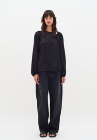 Top lungo in satin nero a maniche lunghe con vestibilità rilassata, caratterizzato da un collo rotondo e dettagli arricciati. Abbinato a pantaloni di denim neri larghi.