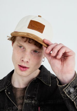 Beige caps med brunt semsket skinnskjerm og et lærlogo-lapp. Bæreren har krøllete hår og er kledd i en mørk denimjakke.