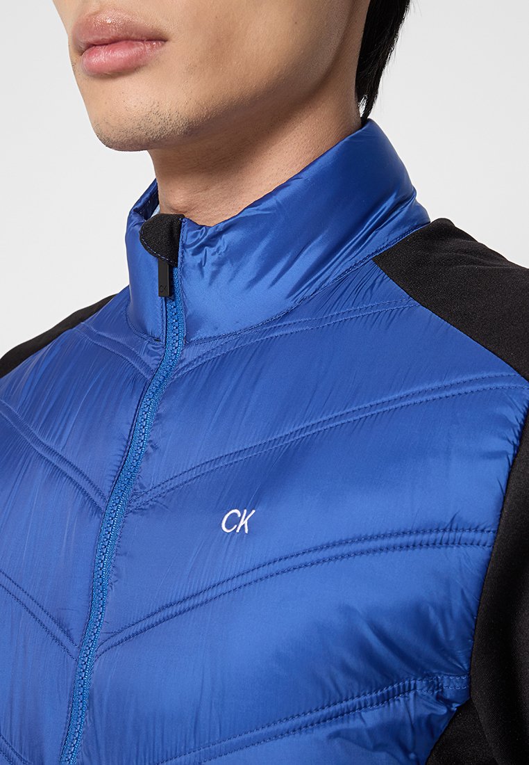 Blauwe nylon jas met een hoge kraag, voorzien van een zwart schouderpaneel en diagonale quiltpatroon. Witte "CK" logo op de borst.