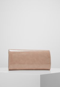 Obdélníková béžová lakovaná kožená kabelka s klopou, umístěná na bílem podstavci před jednobarevným světlým šedým pozadím.