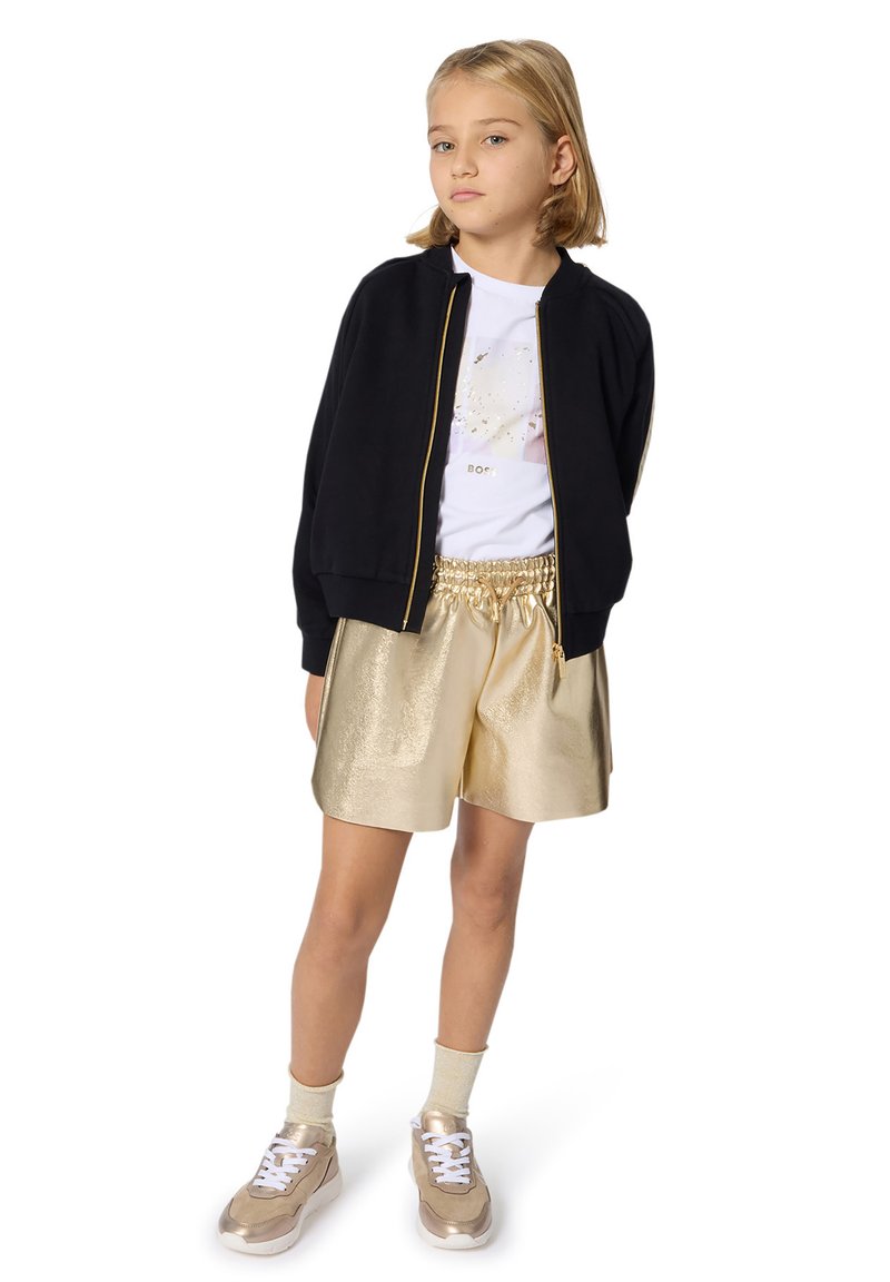 Meisje staat met handen achter haar rug, draagt een zwarte ritssportjack, een witte graphic tee, metalen gouden shorts, crèmekleurige sokken en gouden sneakers.