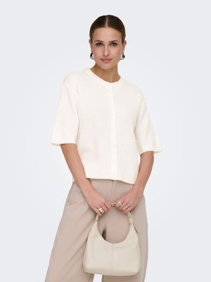 Femme aux cheveux plaqués en arrière, vêtue d'un pull blanc à manches courtes boutonné et d'un pantalon beige, tenant un sac à main beige assorti.