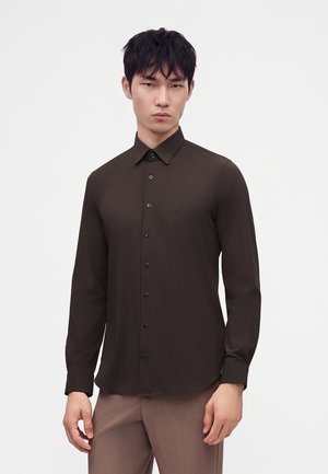 Homme portant une chemise marron foncé ajustée à boutons et un pantalon marron clair, debout devant un fond clair uni.