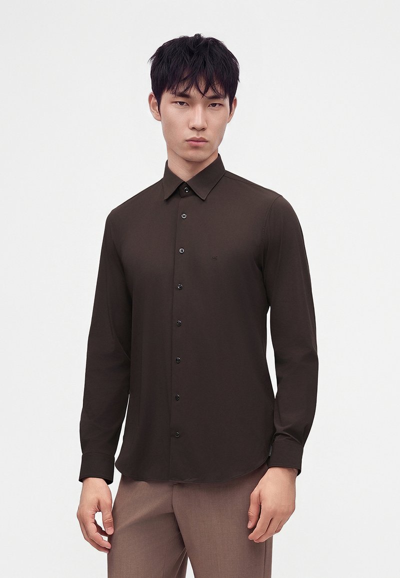 Homme portant une chemise marron foncé ajustée à boutons et un pantalon marron clair, debout devant un fond clair uni.