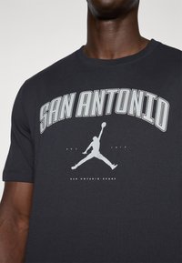Juodas medvilnės marškinėlis su žodžiu "SAN ANTONIO" ryškiomis pilkomis raidėmis virš silueto logotipo, o žemiau užrašyta "EST. 1973" ir "SAN ANTONIO SPURS".