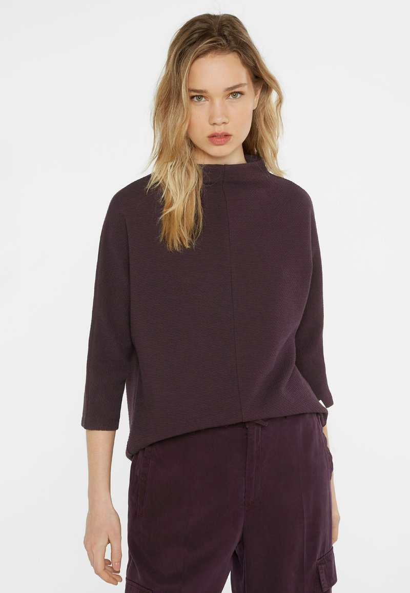 Lila strukturierte Bluse mit Stehkragen, Dreiviertelärmeln und entspannter Passform, getragen mit passenden tieflila Hosen.