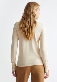 Maglione beige a maniche lunghe in morbido tessuto a maglia, con una forma aderente e dettagli con bottoni sui polsini, indossato con pantaloni marroni.
