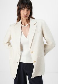 Kremfarget blazer i strukturert stoff med ensidig design, to frontlommer og gullfargede knapper. Hvit topp med V-hals under.
