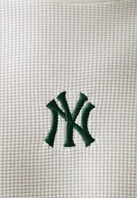 Logo intrecciato "NY" ricamato in verde su sfondo di tessuto bianco dalla texture evidente.