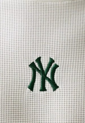 Logotipo "NY" entrelazado verde bordado sobre fondo de tela blanca texturizada.
